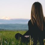 Woman enjoying peace in a lush green field with a scenic mountain view during sunset.