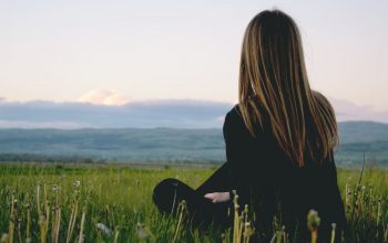 Woman enjoying peace in a lush green field with a scenic mountain view during sunset.